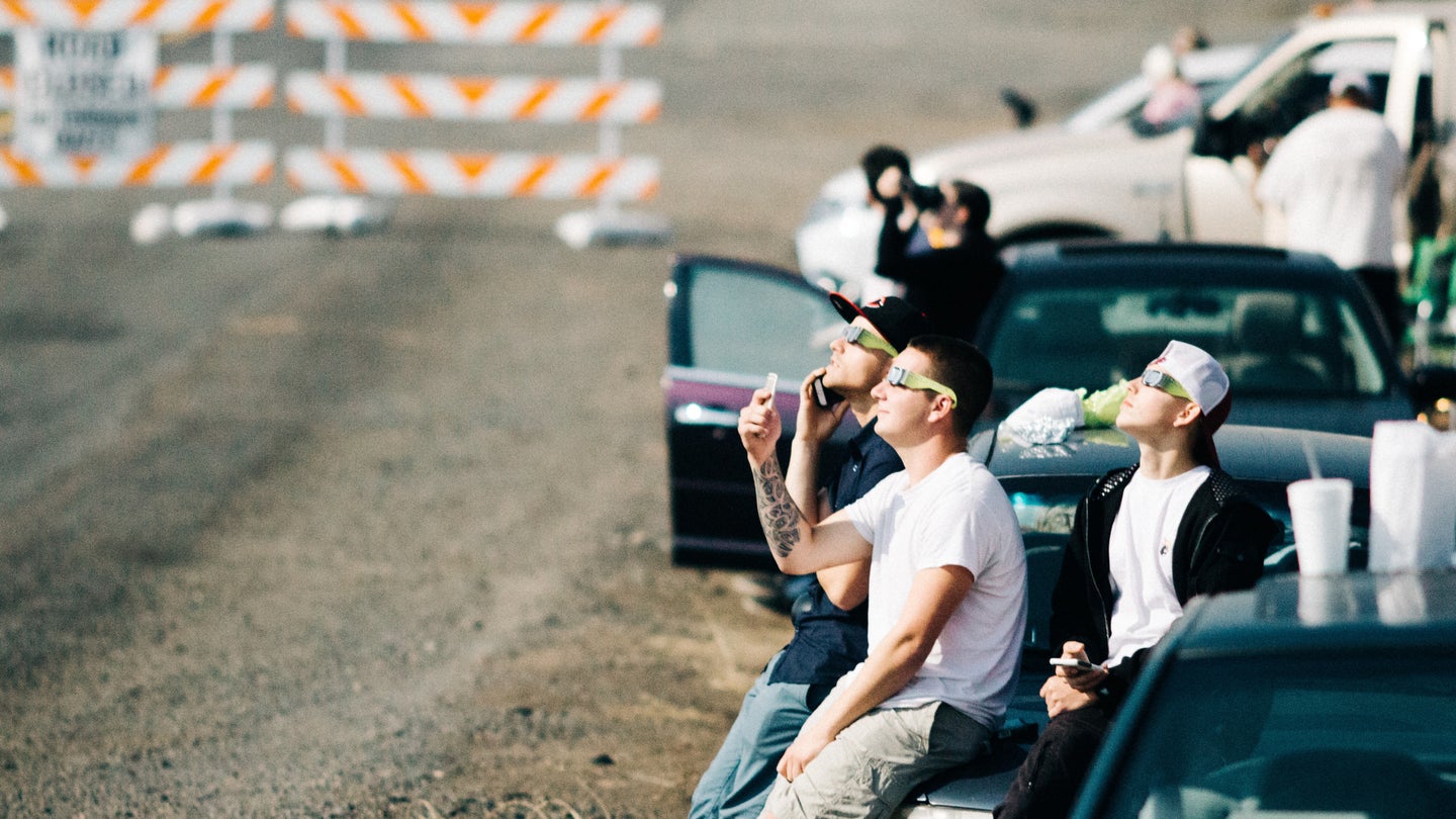 A group of people outside their cars along a closed road, wearing eclipse glasses to watch a solar eclipse.