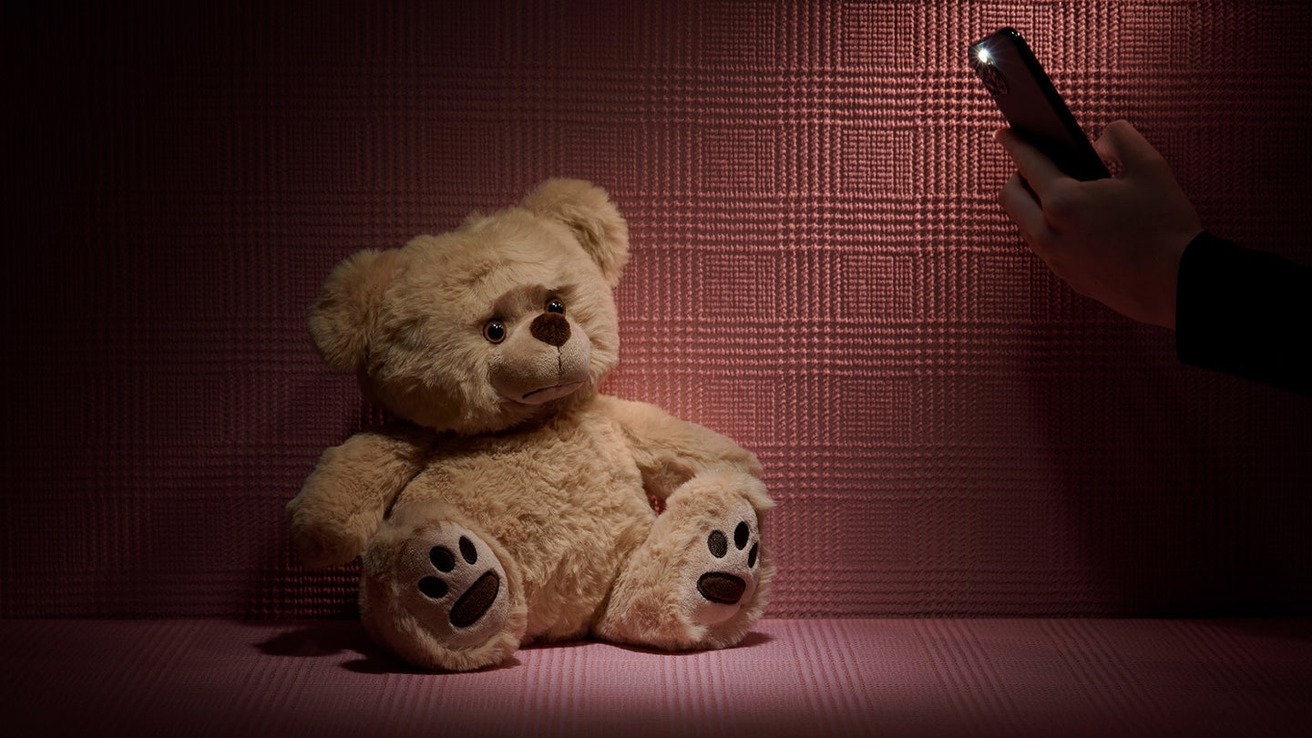 Teddy bear sitting in a corner and hands peeking from the corner with a phone taking pictures of the teddy bear.