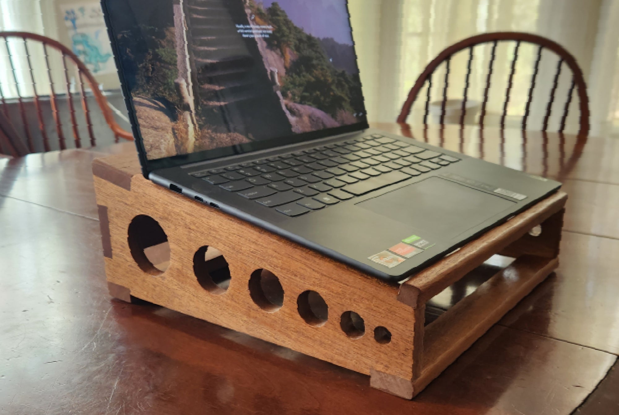 A DIY wooden laptop stand on a wooden table in a dining room, with a laptop on it. The rabbet joints in the side panels are visible.