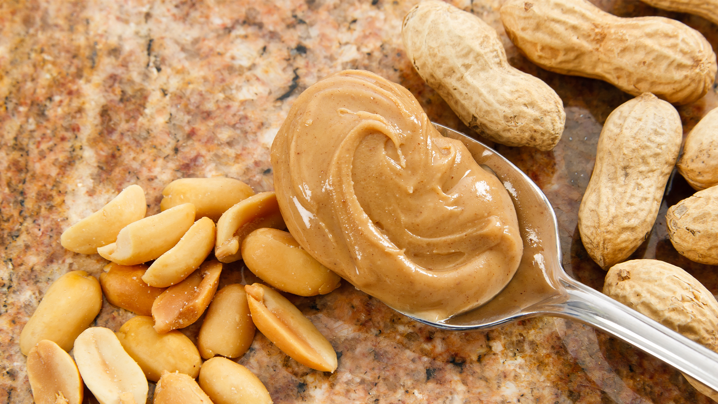 A spoon of creamy peanut butter over a slice of bread and surrounded by peanuts, shelled and unshelled.