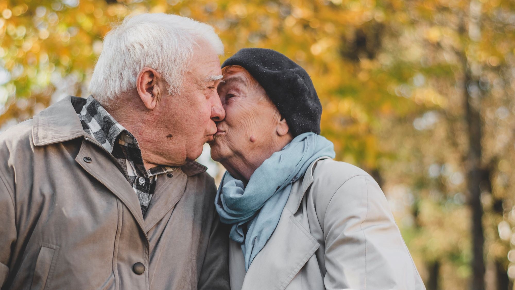 An older couple shares a kiss against a backdrop of fall trees.