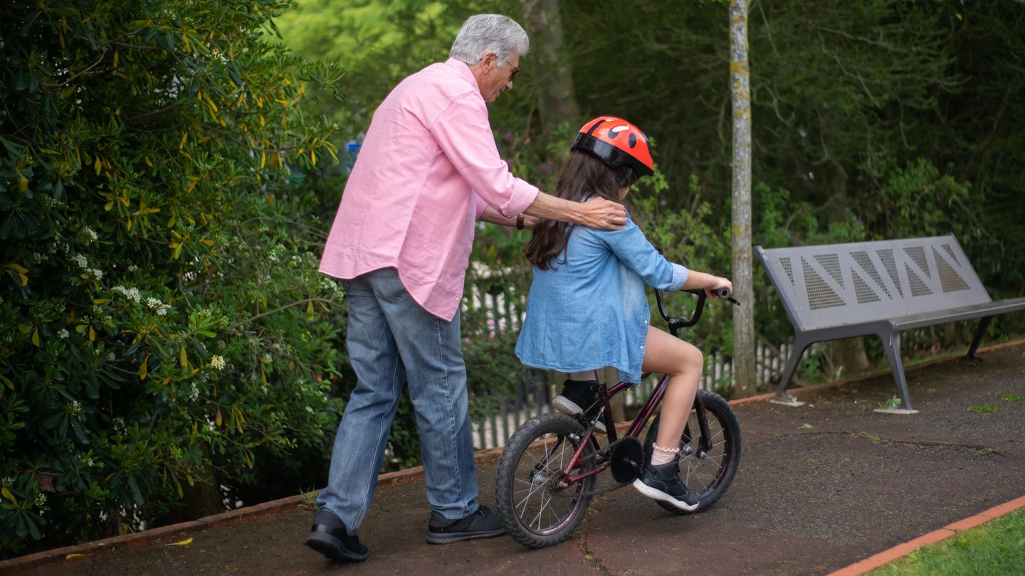 An elderly person with short gray hair, wearing a pink shirt and jeans, with their hands on the shoulders of a kid they're teaching how to ride a bike. The child is wearing a red helmet and using a balance bike on a park path.