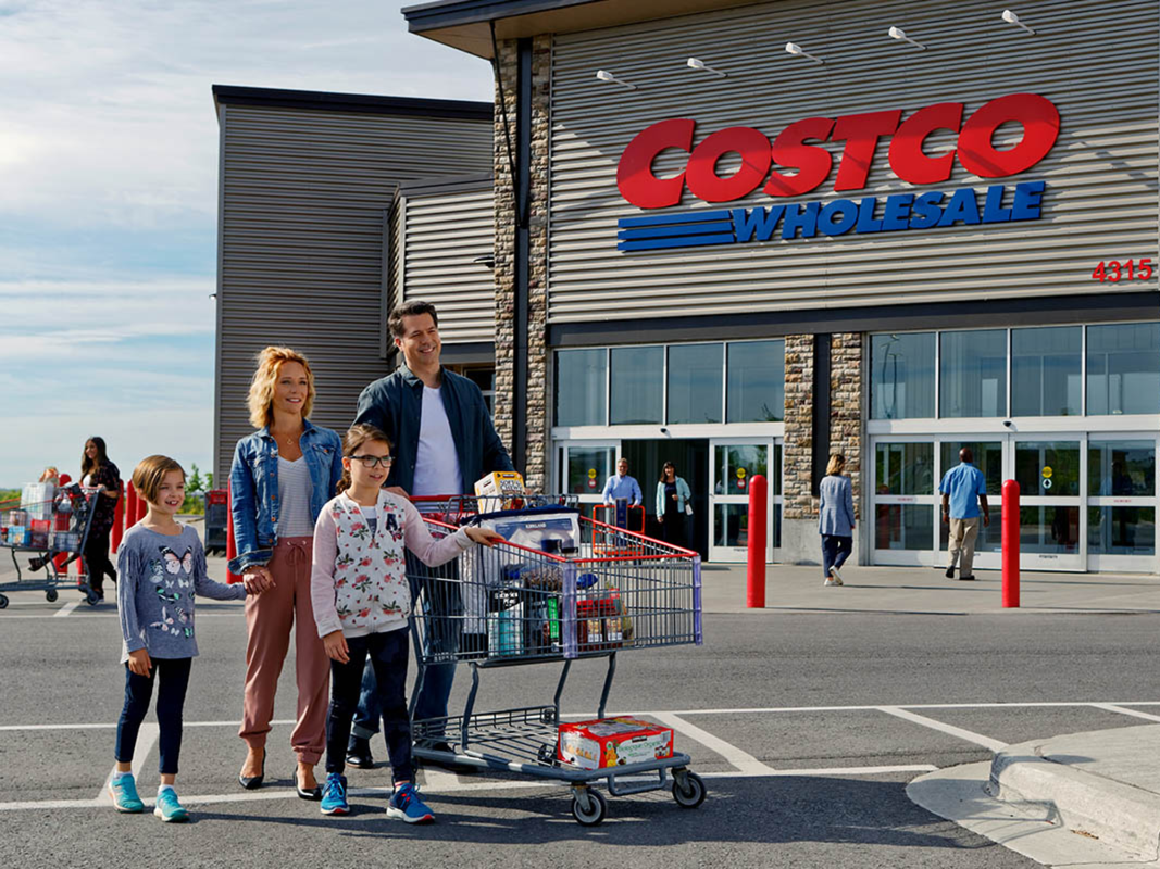 A family walking out into a Costco parking lot with a cart filled with groceries