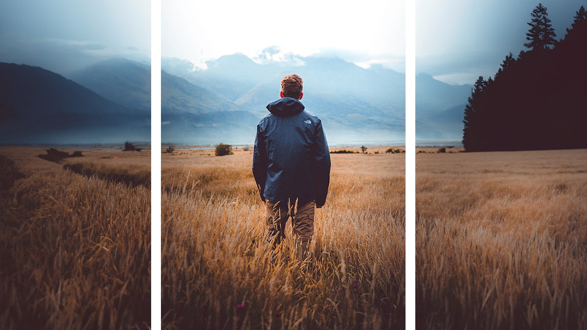 A man in a blue jacket standing in a golden field of grass, looking at blue foggy mountains in the distance, with more of the landscape added on the left and right using Adobe Photoshop's AI image extender tool, Generative Fill.
