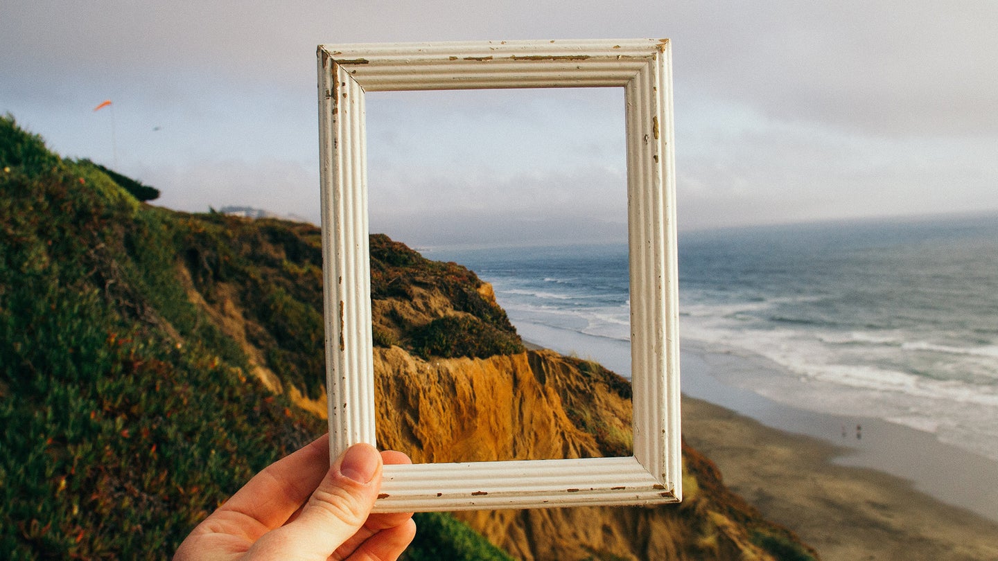 A person holding an empty photo frame up against a seaside cliff landscape, showing how an AI image extender can add pixels beyond the edge of an original photo.