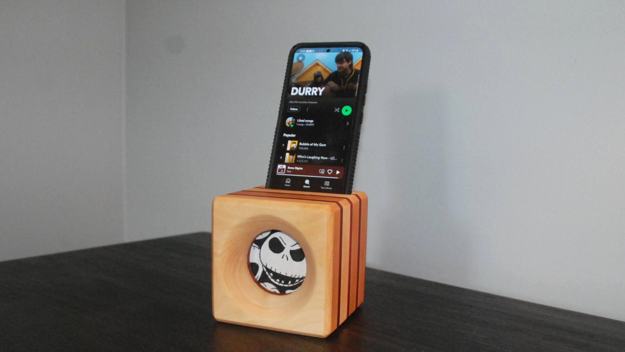 A pale beige DIY wooden phone amplifier with a Jack Skellington fabric speaker cover on a black shelf against a white wall, with an iPhone inserted into the top, playing music from the band Durry.