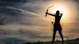 The silhouette of a female hunter with a bow and arrow at sunrise.