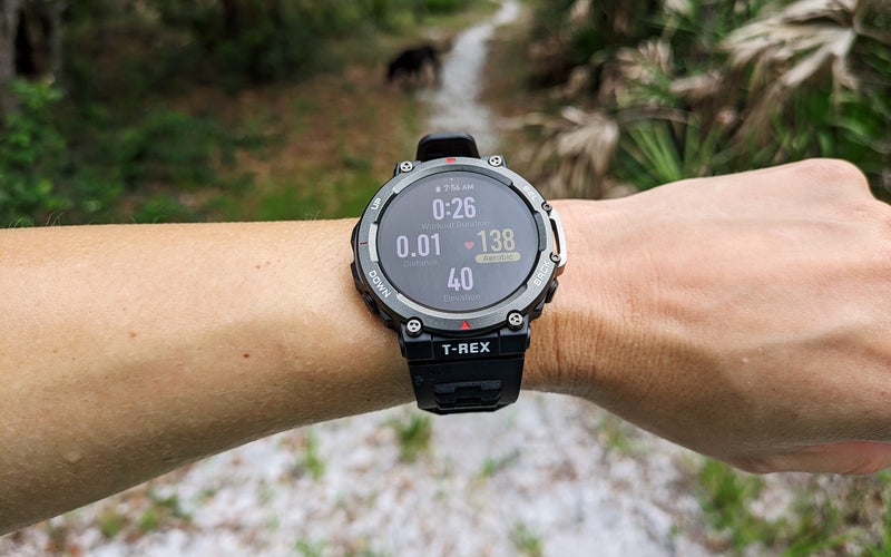 Amazfit T-Rex 2 hiking watch on a wrist in front of a trail through the woods
