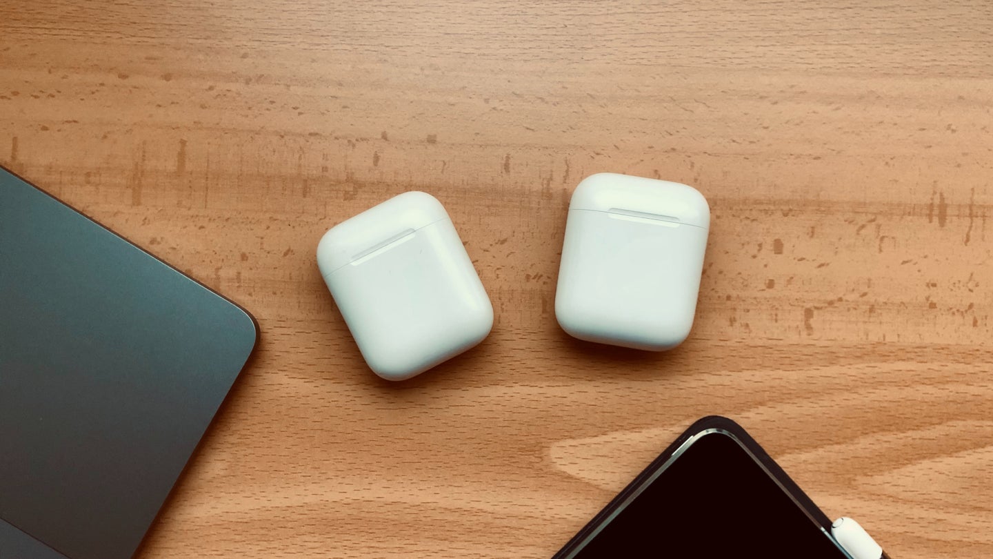 Two pairs of AirPods on a wooden table next to a black iPhone and a Macbook.