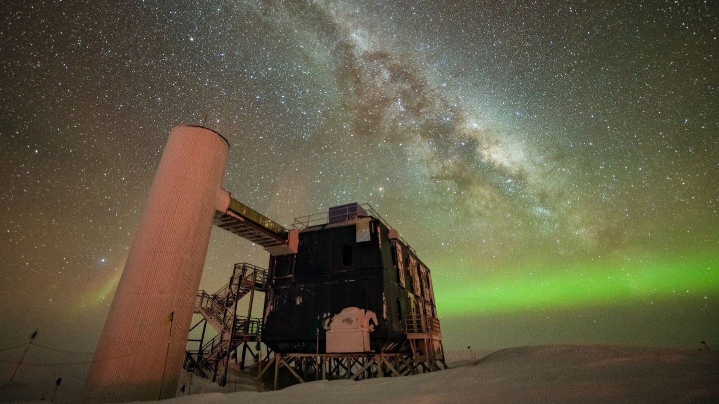 The IceCube Lab is seen under a starry night sky, with the Milky Way appearing over low auroras in the background.