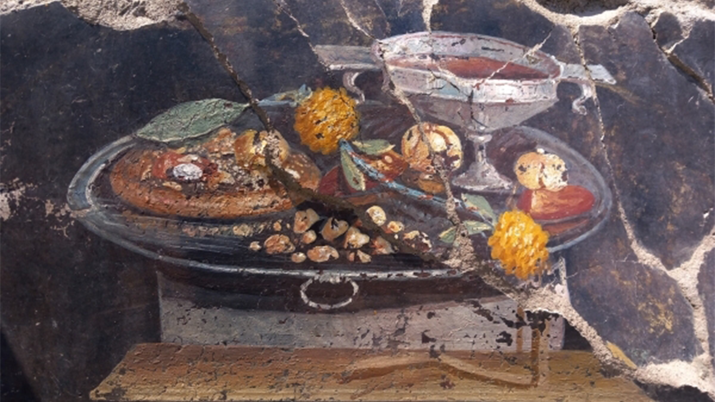 A fresco painted on a wall in the ancient city of Pompeii that depicts a focaccia with fruit and possibly pesto sauce on it. It is served on a silver tray and has a wine chalice next to it.