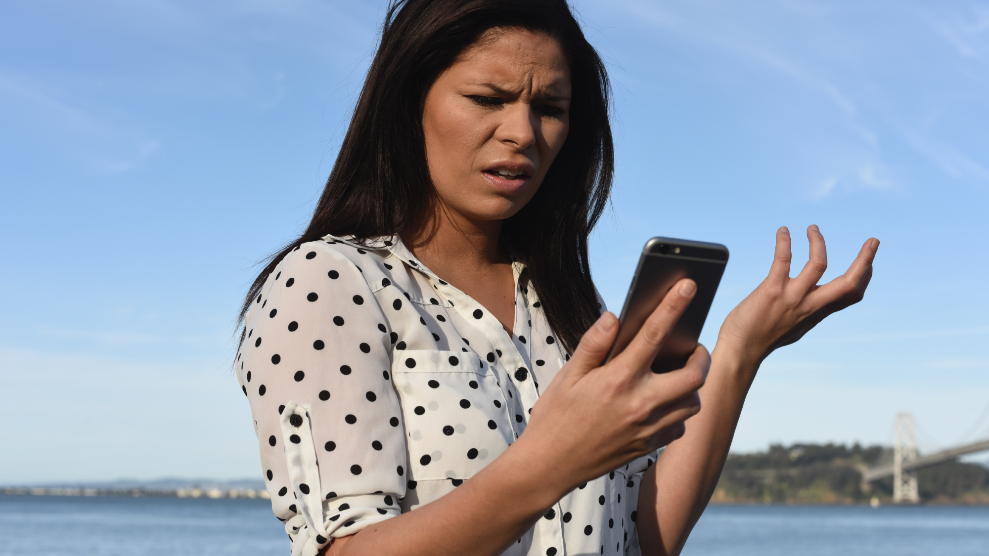 A woman with black hair wearing a white shirt and standing in front of a body of water while looking very confused at her phone.