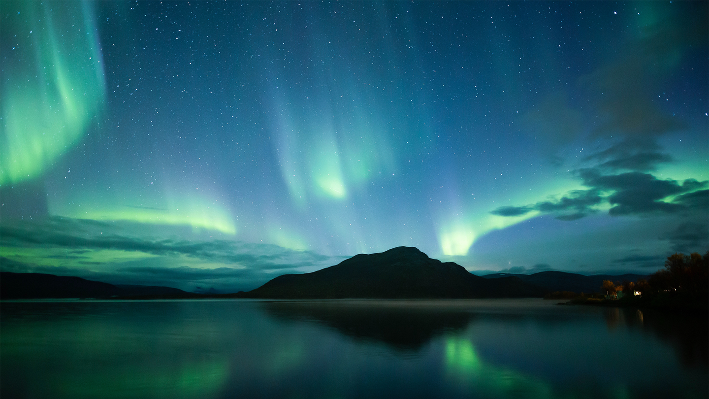A green and blue aurora borealis glows above a body of water and mountains.