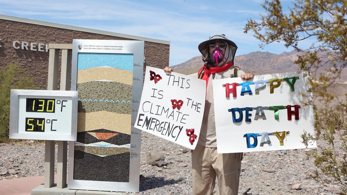 A protester stands next to a digital display of an unofficial heat reading at Furnace Creek Visitor Center during a heat wave in Death Valley National Park in Death Valley, California, on July 16, 2023. The signs read “this is the climate emergency,” and “happy death day.”