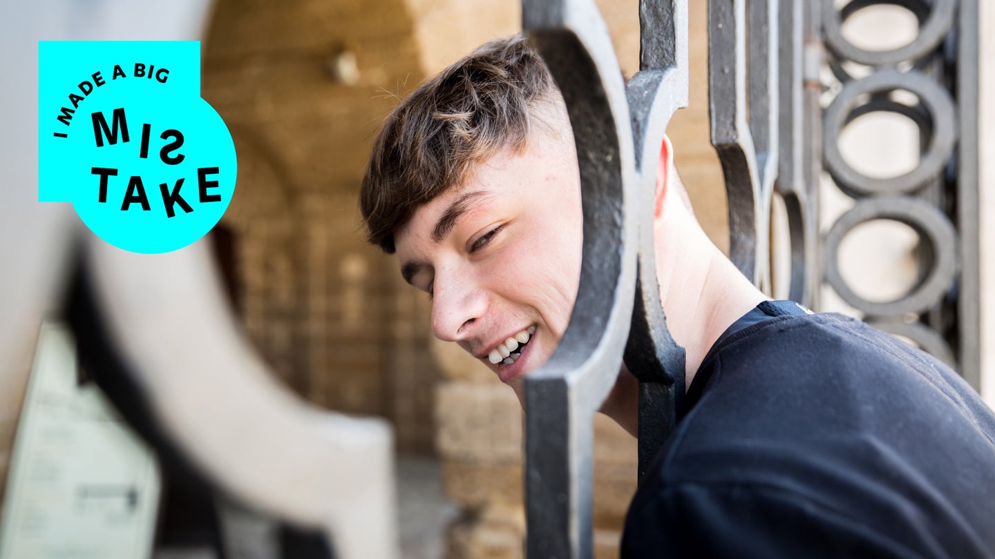 A short-haired teenage boy wearing a black shirt and grinning with his head stuck in a metal fence.