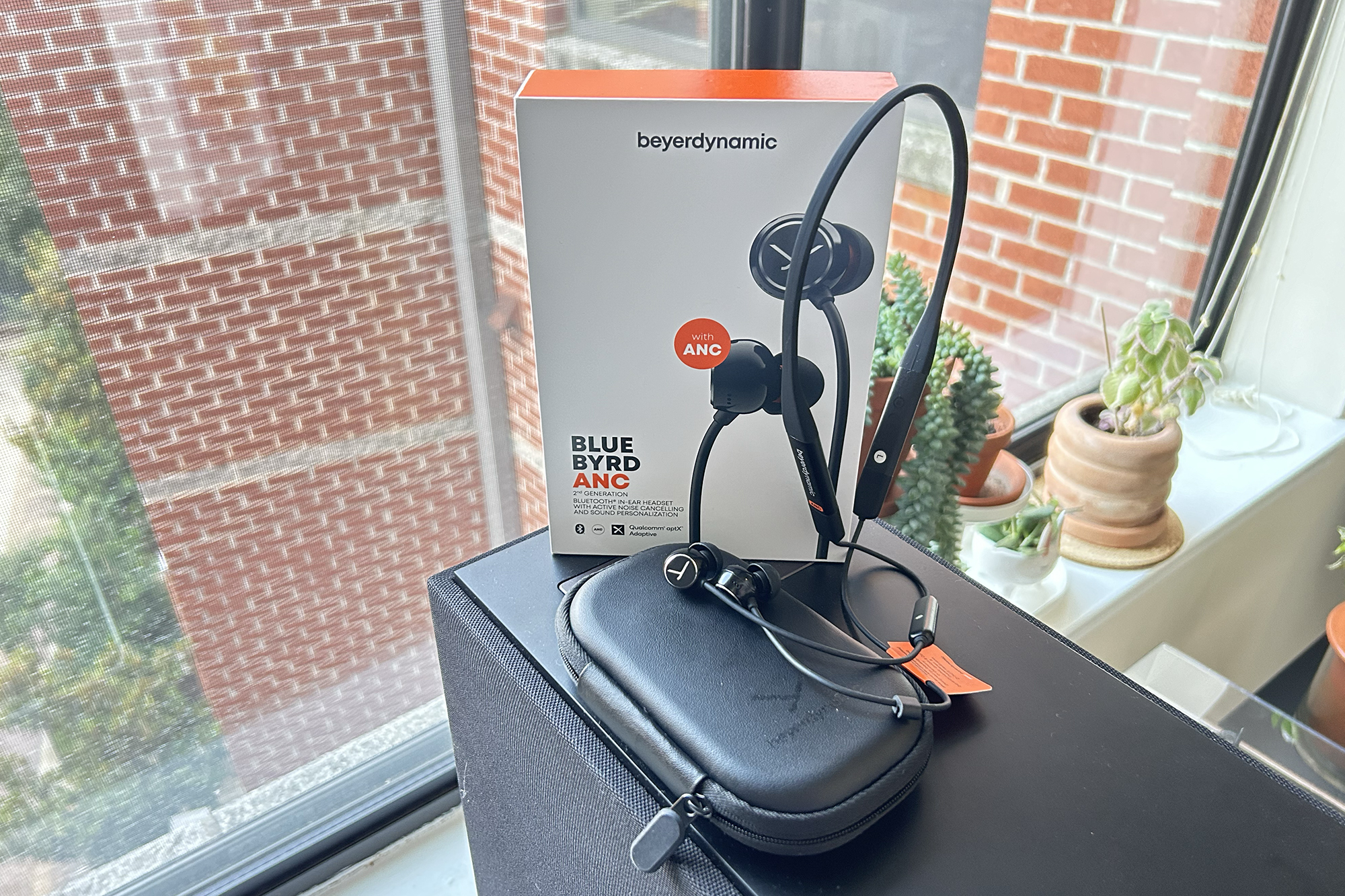 beyerdynamic Blue BYRD ANC neckband earbuds, box, and case sitting on a speaker in front of a window