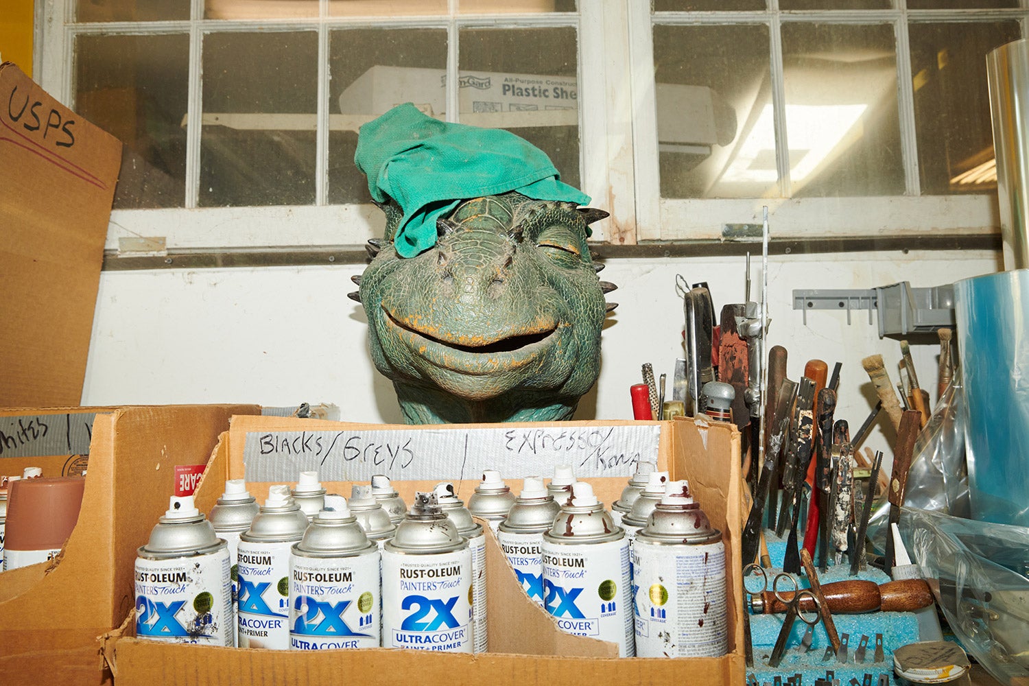 head of dinosaur sculpture sits behind pray paint and next to tools in workshop storage area.