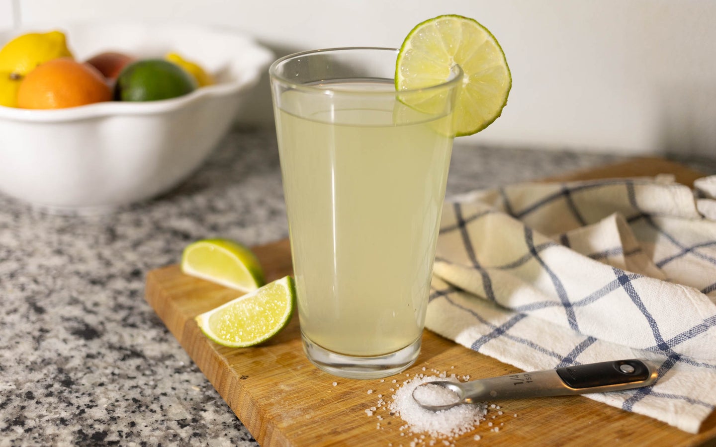 A homemade electrolyte drink that's a pale green lime color, on a cutting board next to some lime slices and sugar.