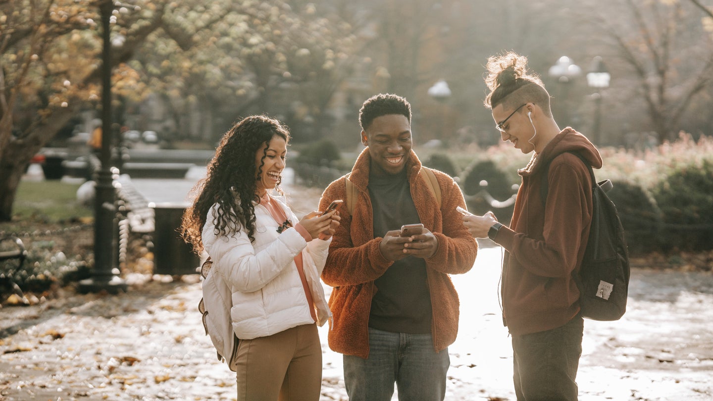 Three young people of different races and genders standing on a sidewalk in a park during the fall, all holding their phones and laughing.