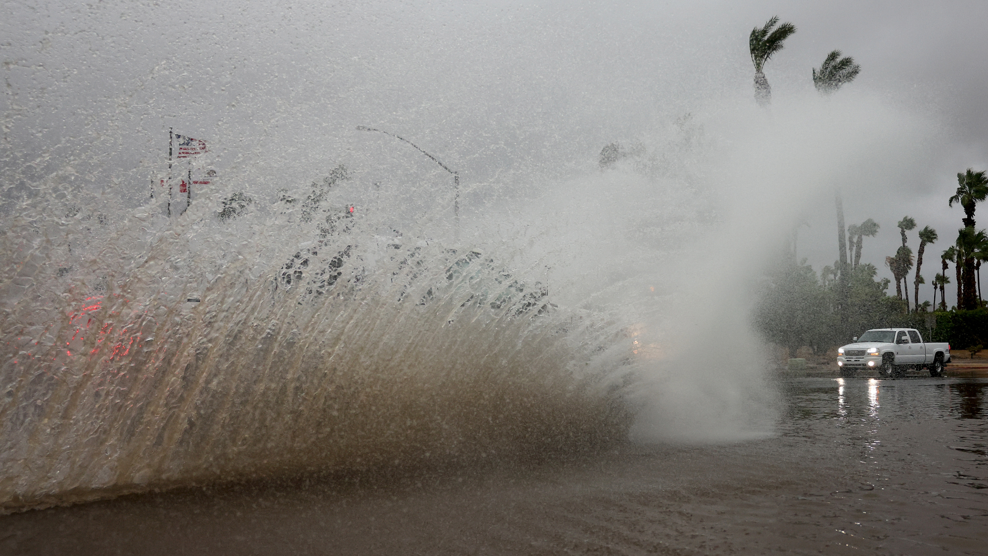 A vehicle drives through a flooded street as Tropical Storm Hilary moves through the area on August 20, 2023 in Cathedral City, California. A wave of water pushes onto the street and palm trees shake in the wind.