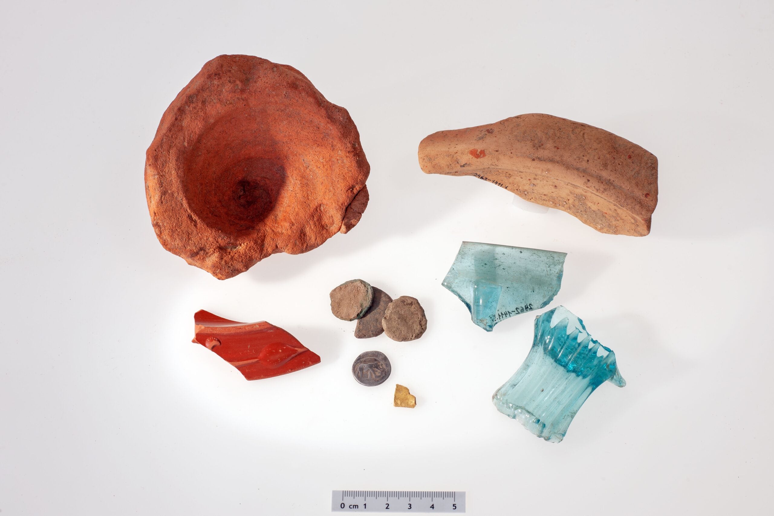 Small selection of Roman finds (from top left to bottom right): An amphora base, the shard of a mortar, the rim of a small bowl of Roman tableware with a red coating (terra sigillata), four coins in as-found condition, one of which was silver from Julius Caesar, Fragment of a gold object, pieces of a square bottle and a blue glass ribbed bowl. CREDIT: ADA Zug, Res Eichenberger