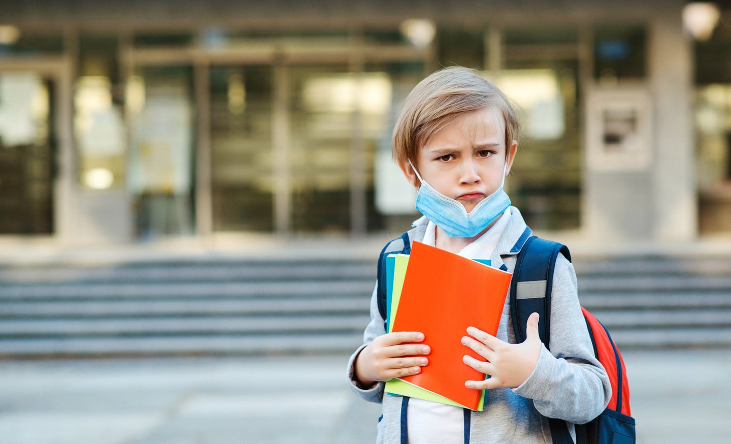 Kid with blonde hair wearing a blue COVID mask holding books and a backpack on the first day of school