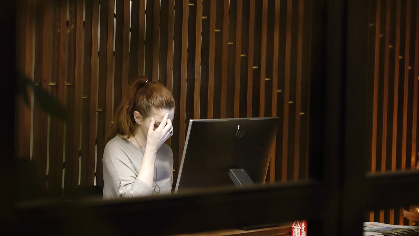 A woman sitting in front of a Mac desktop computer with her hand on her face, viewed through a window from outside.