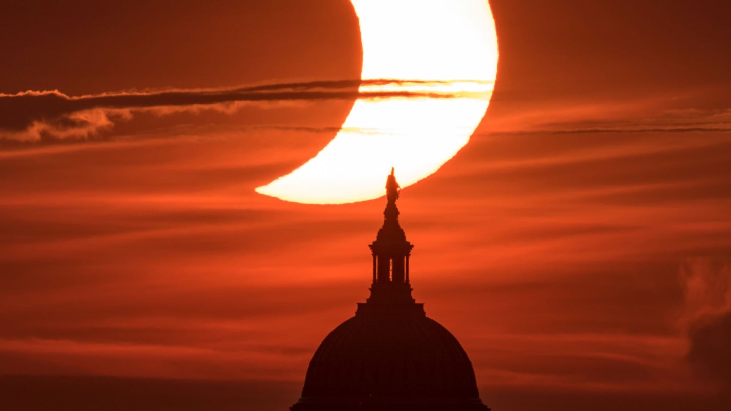 A yellow sun partially blocked by a moon in a reddish sky with the US capitol in the foreground.