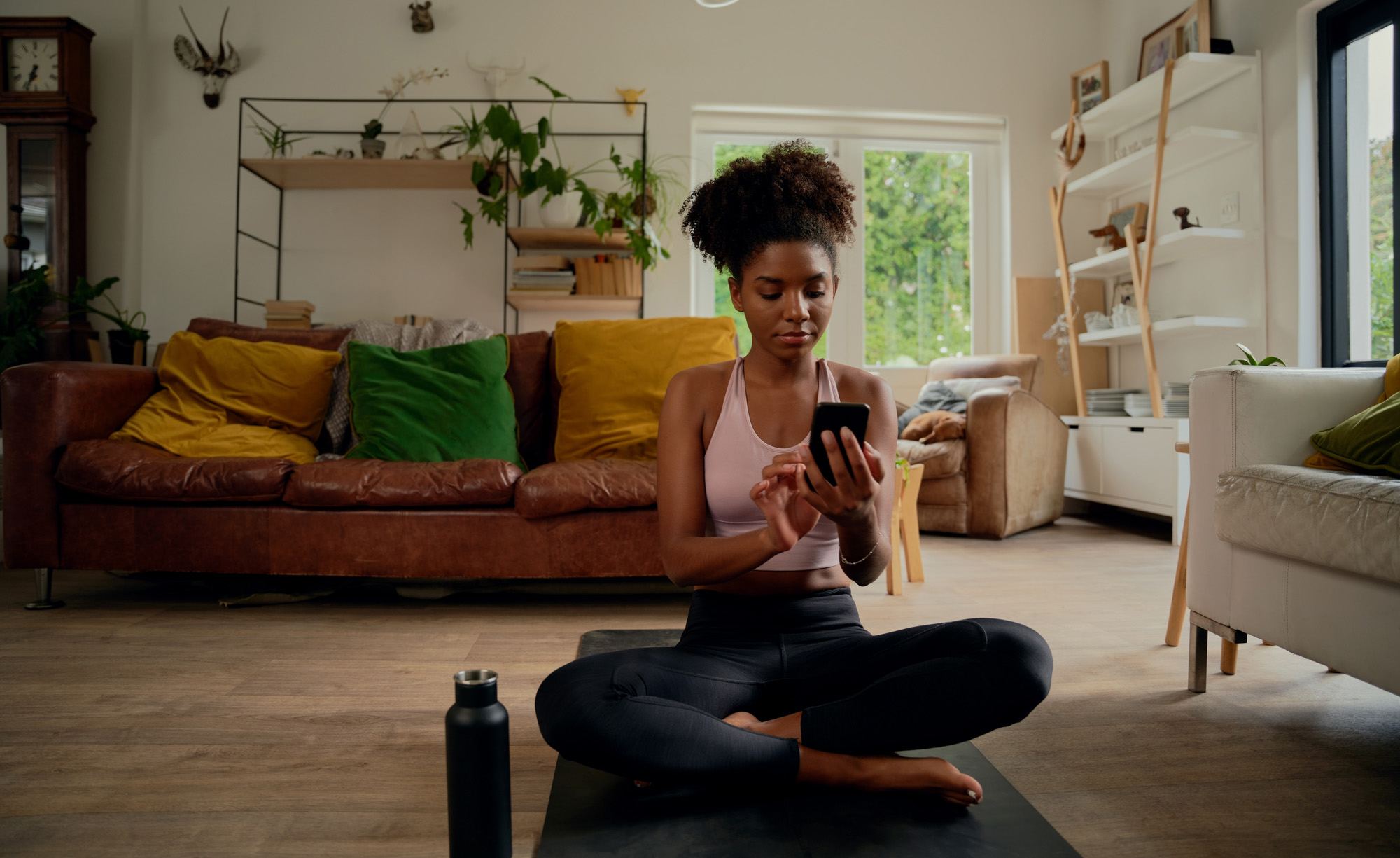 A woman sitting in her living room looking at her smartphone while sitting on her yoga mat.