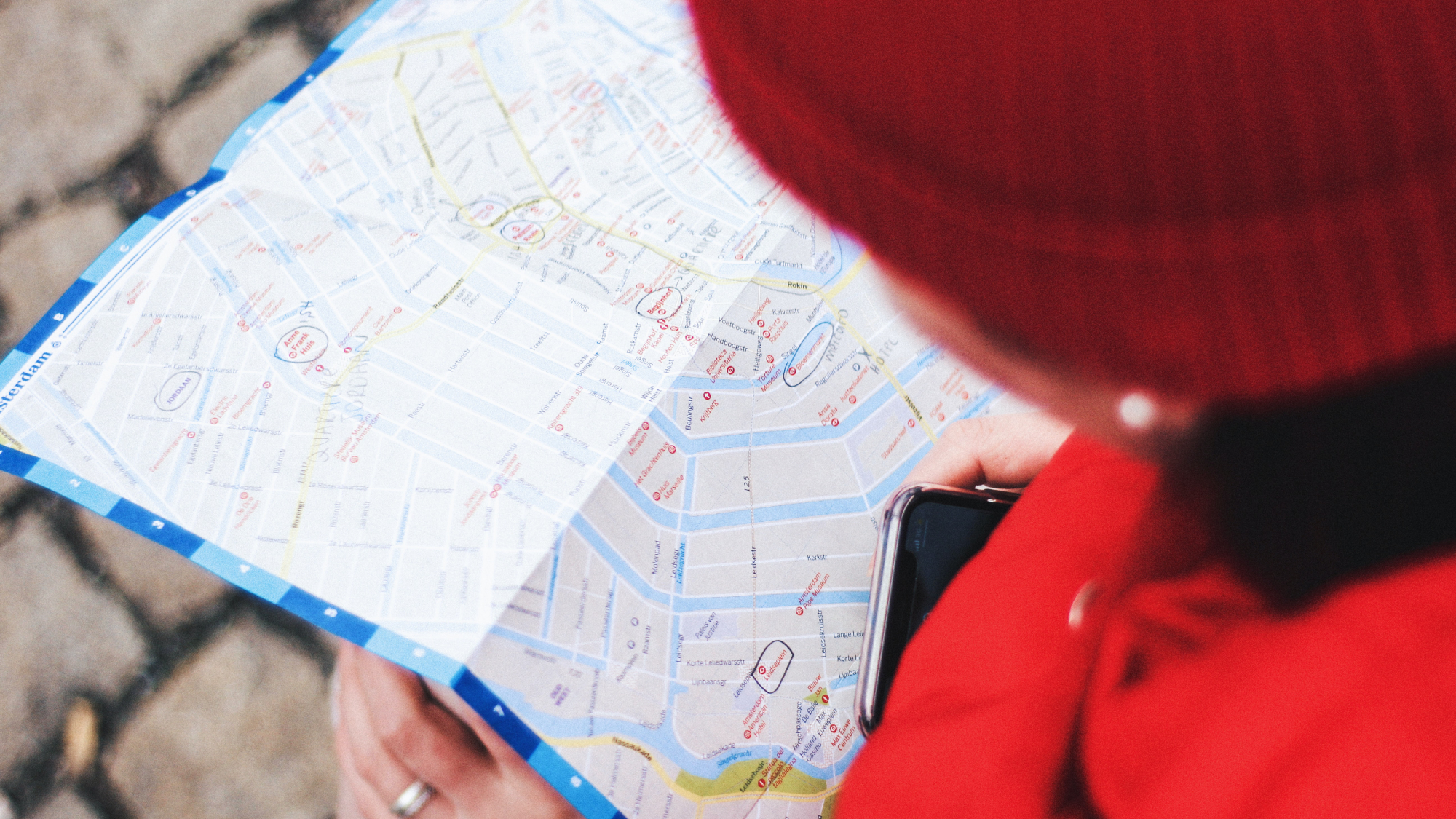 A person wearing a red jacket and a red knit beanie, looking at a paper map while holding their phone, possibly as they prepare to share their location with someone else via a location-sharing app.