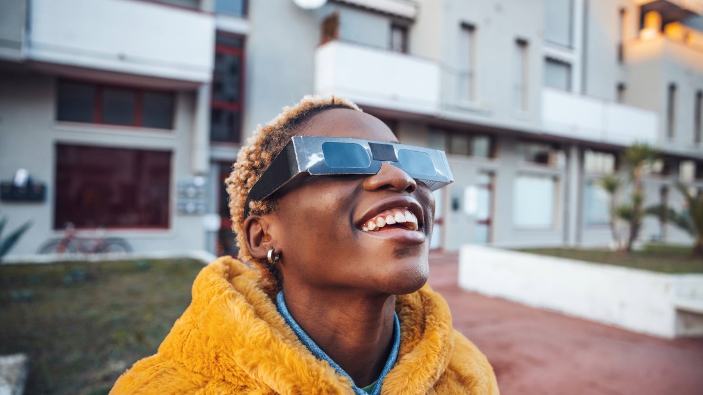 A young Black person with short dyed-blonde hair wearing a yellow jacket and eclipse glasses while looking up at an eclipse in front of some residential buildings.