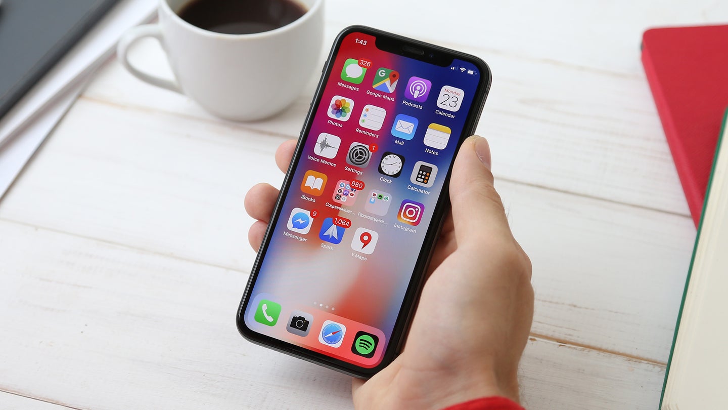 A person holding an iPhone with a bunch of apps on the home screen. A cup of coffee is behind their hand on a white table.