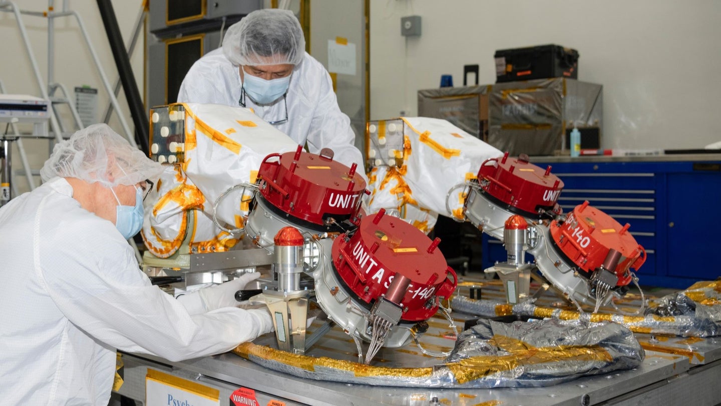 Red-capped Hall effect thrusters being attached to a spacecraft body by two engineers.