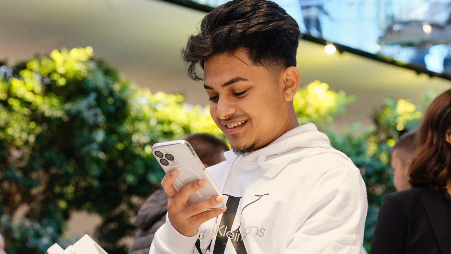 A man wearing a white hoodie stands outside and smiles as he looks as his iPhone 15