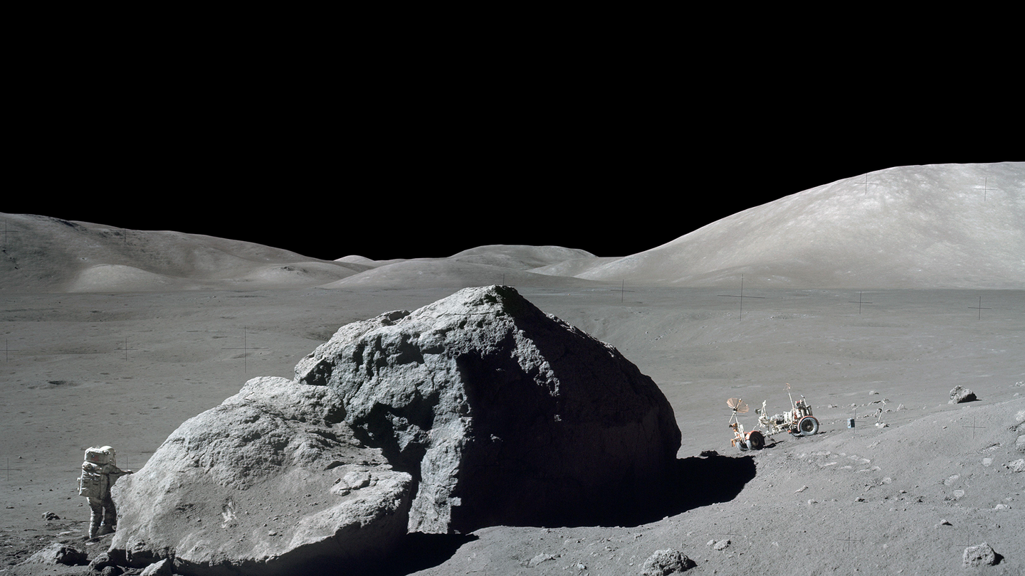NASA astronaut Harrison Schmitt is standing on the moon’s surface next to a large split lunar boulder, with a roving vehicle on the other side of the boulder.