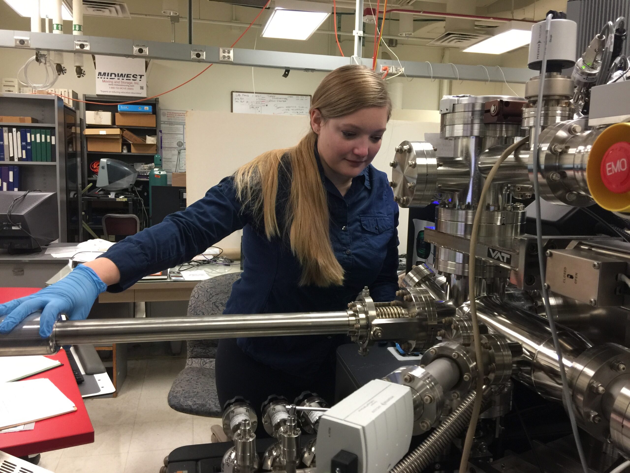 A scientist named Jennika Greer works with an atom probe in a lab. She is holding a long, metal tube and looking towards a special microscope.