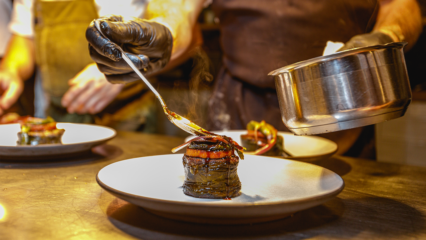 A steak with kelp on top is plated by a chef holding a sauce pan.