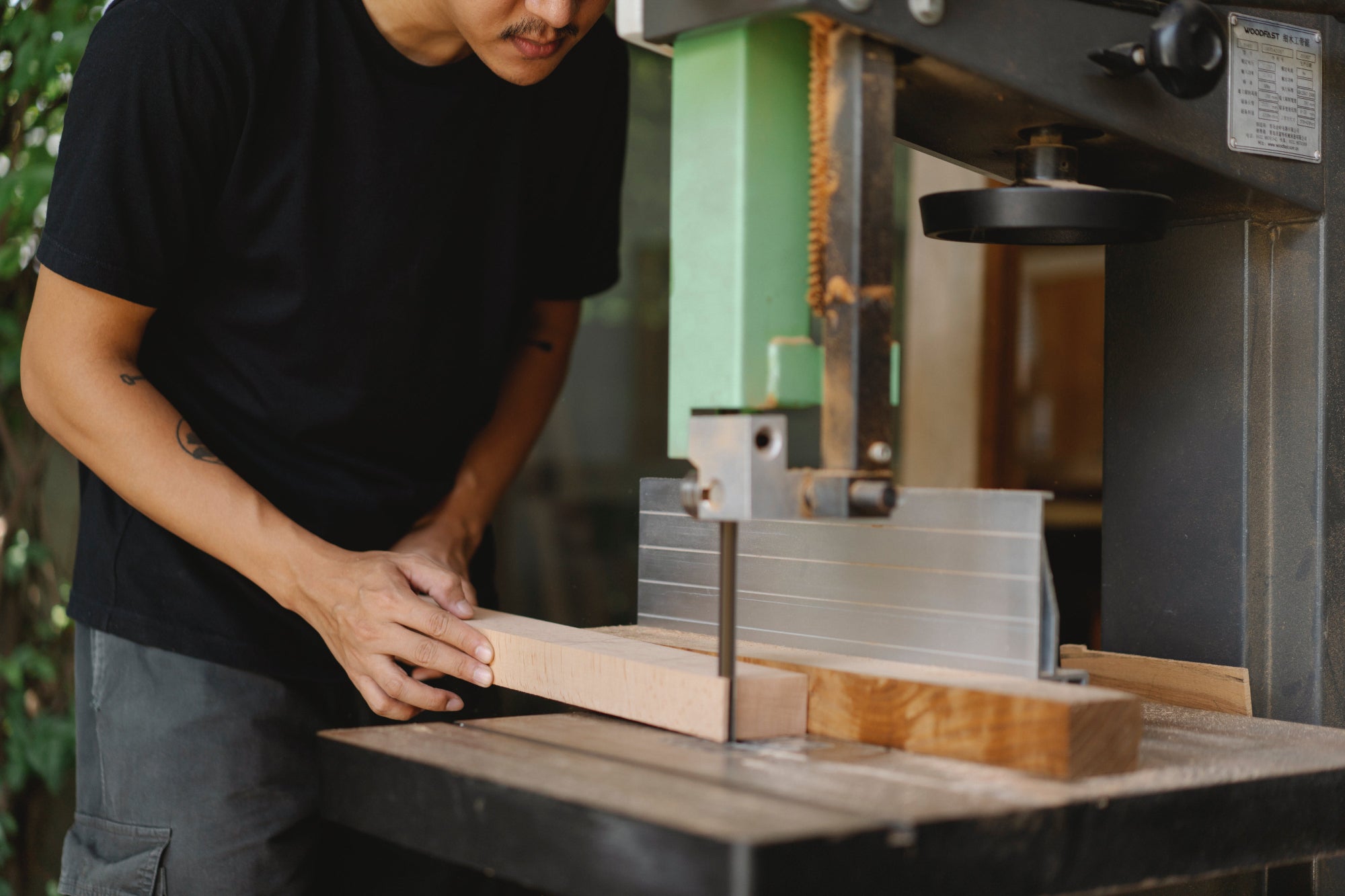 A person wearing a black t-shirt using a band saw to cut a piece of wood in a woodshop.