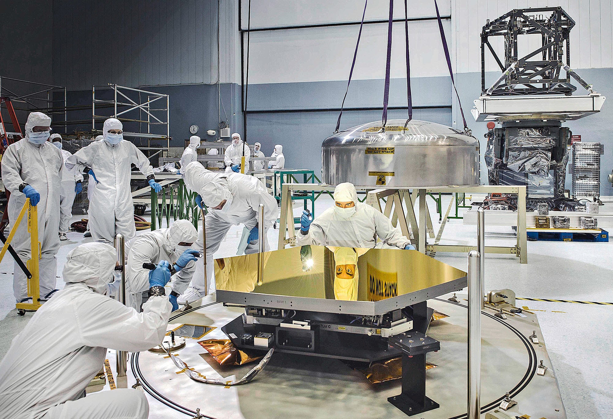 A team of scientists in a large room examines a golden mirror for the James Webb Space Telescope.