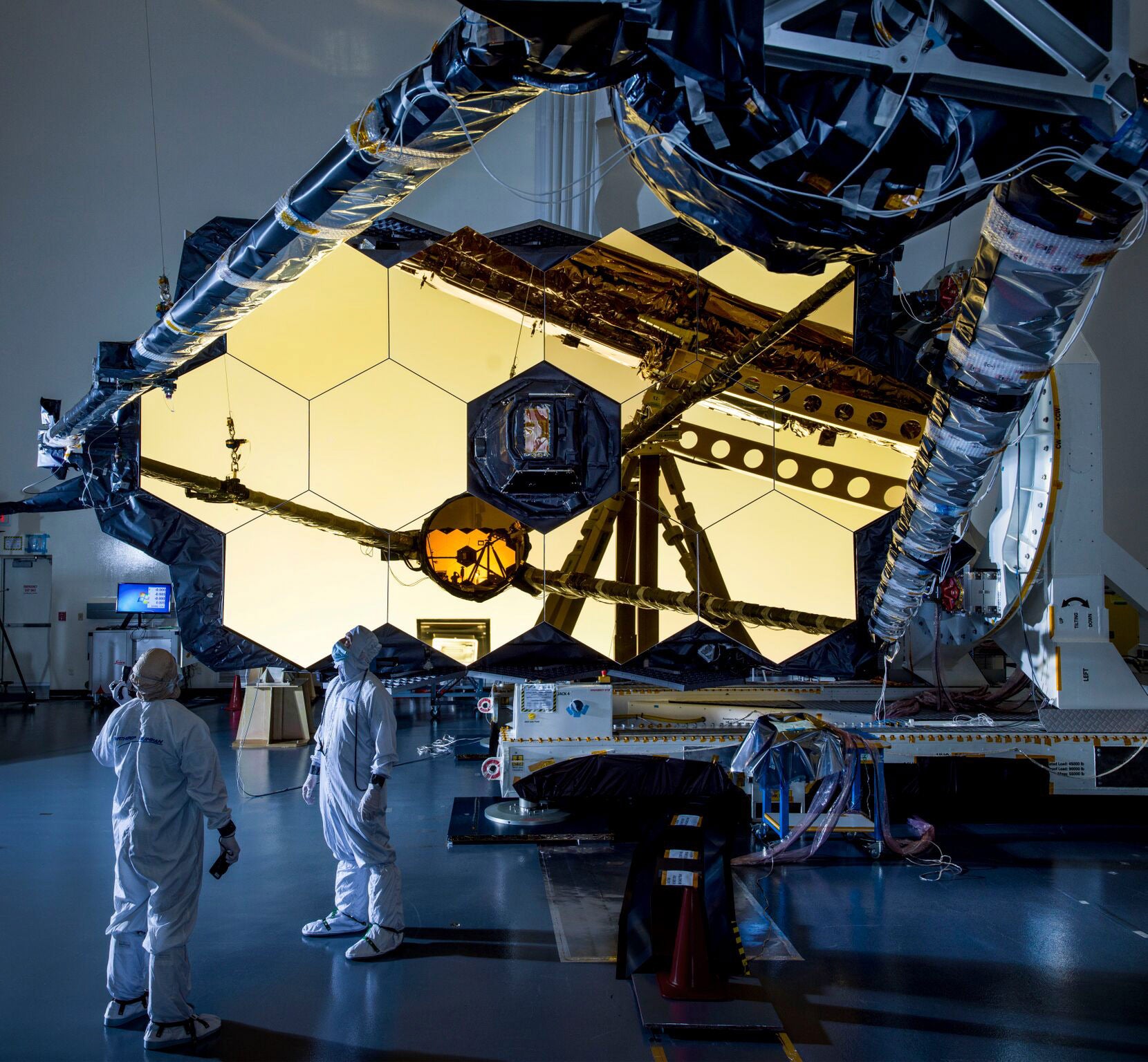 Two scientists stand in front of the golden JWST mirrors in a large, dimly lit room.