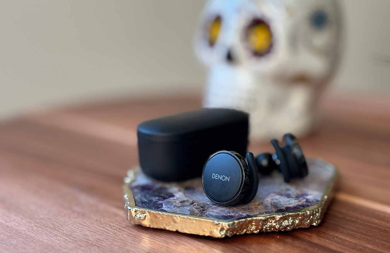 Black matte Denon PerL Pro True Wireless earbuds on an amethyst coaster in front of a colorful sugar skull candle