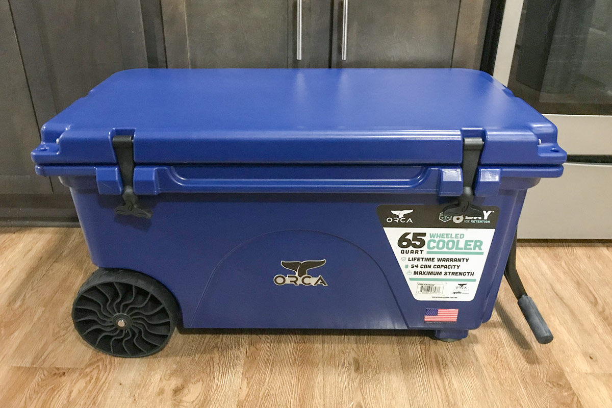 A blue ORCA 65-Quart Insulated Rolling Cooler sits on the floor in front of some kitchen cabinets.