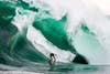 A surfer makes his way across a massive, irregularly shaped, emerald-green wave.