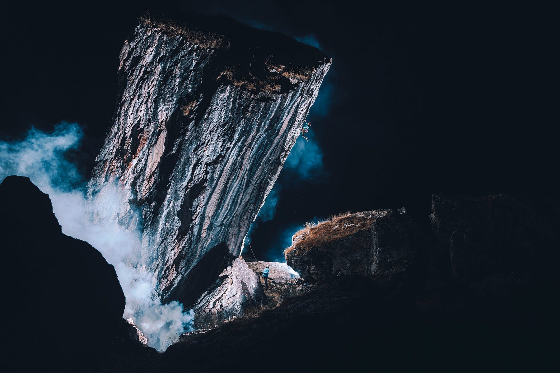 A climber scales a large monolith sticking out of the ground at night, with smoke shrouding the bottom. 