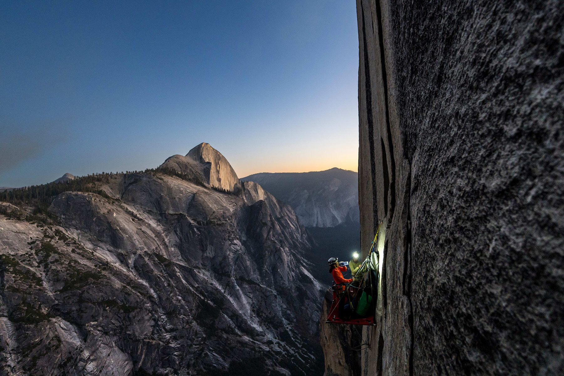 Two climbers with headlamps hang from the face of a cliff in Yosemite with mountains in the Yosemite Valley in the background.