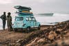 Two people stand beside a retro-looking blue car stacked high with surfboards while looking at an ocean swell.