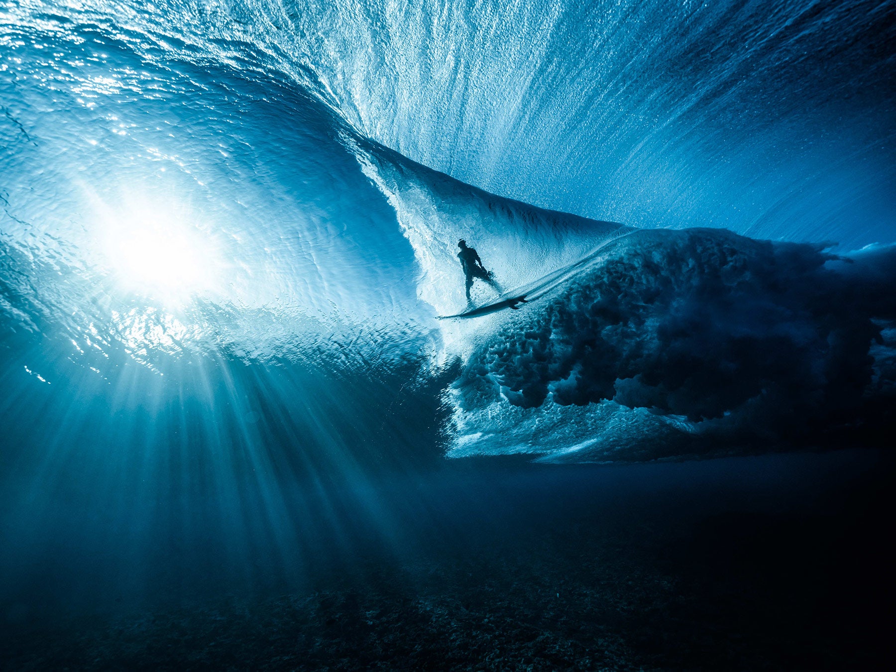 A surfer is seen from beneath the wave while rays of sunlight cut through the darkness.