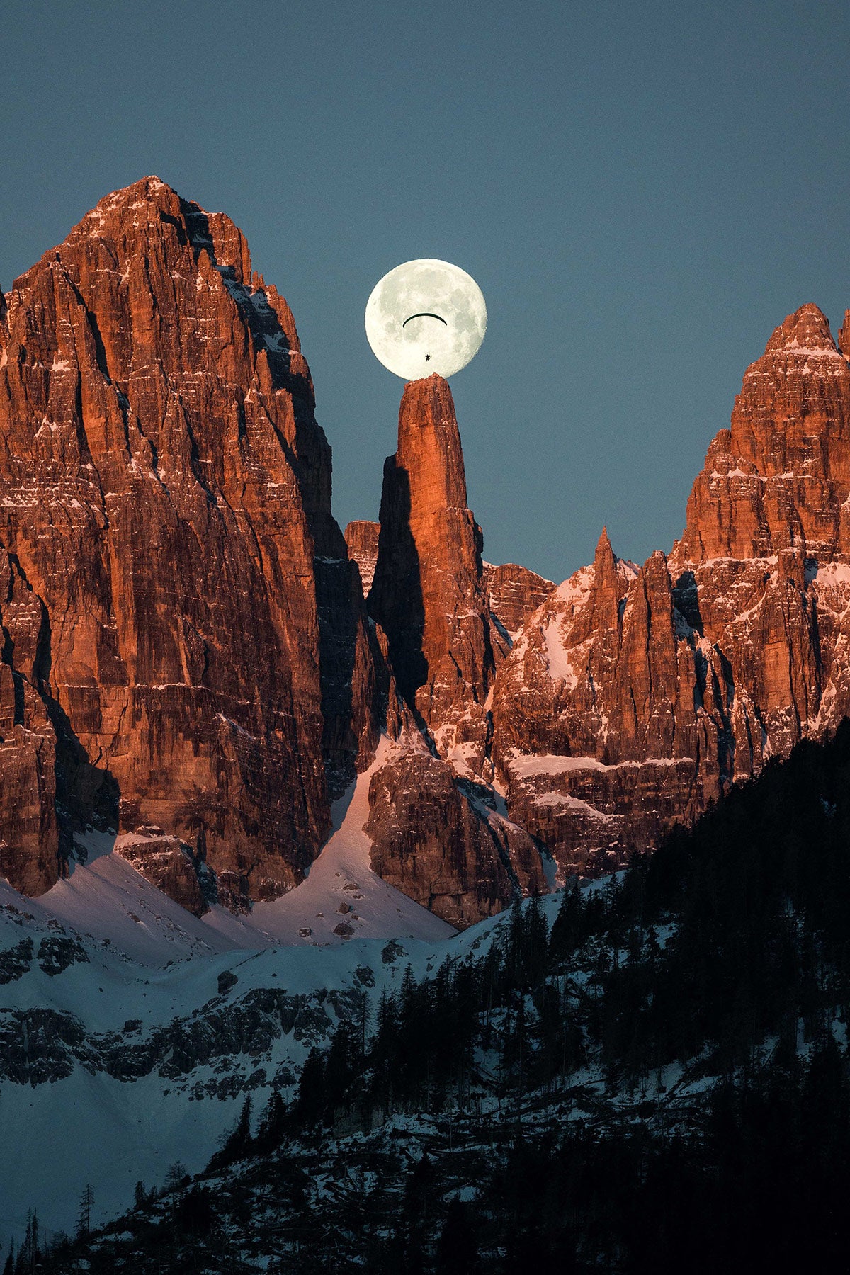 Steep red mountains stick out of the snow-covered ground with a full moon perfectly placed at the top of one spire.