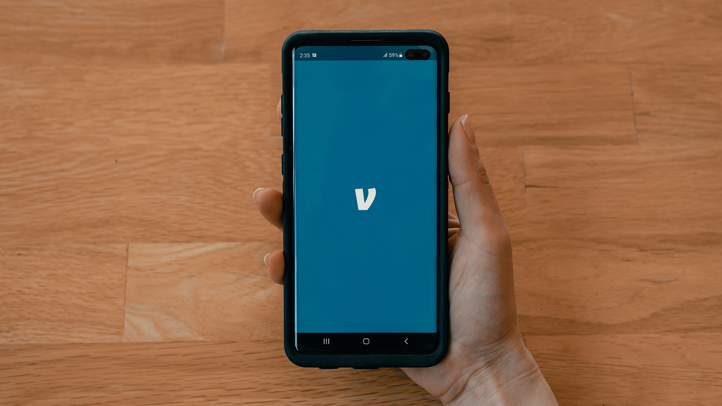 A hand holding a phone over a wooden table. The phone screen is blue and shoes the logo for the Venmo app.
