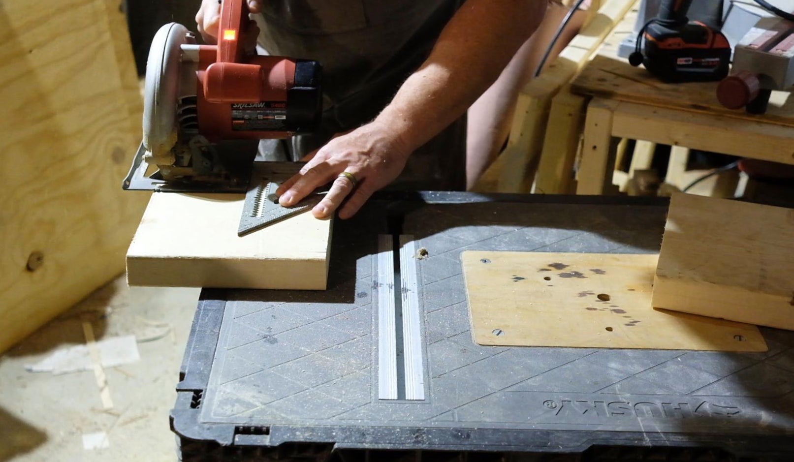 A man in a workshop uses a circular saw and speed square to make a square cut on a small piece of wood.